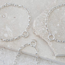 Three silver chain bracelets with one central ring detail, shown on a grey stone