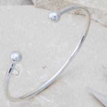 A silver open cuff with silver orbs on the open ends, displayed upright leaning against beige stone.